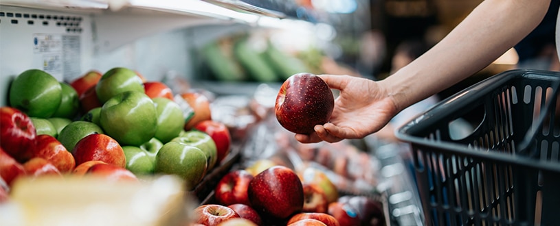 Person holding a shopping basket as they are picking out an apple from a wide variety of fruits at the grocery store.