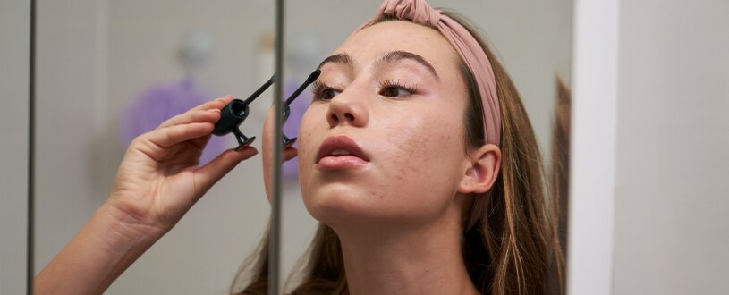 Young woman looking in bathroom mirror while applying drugstore mascara.